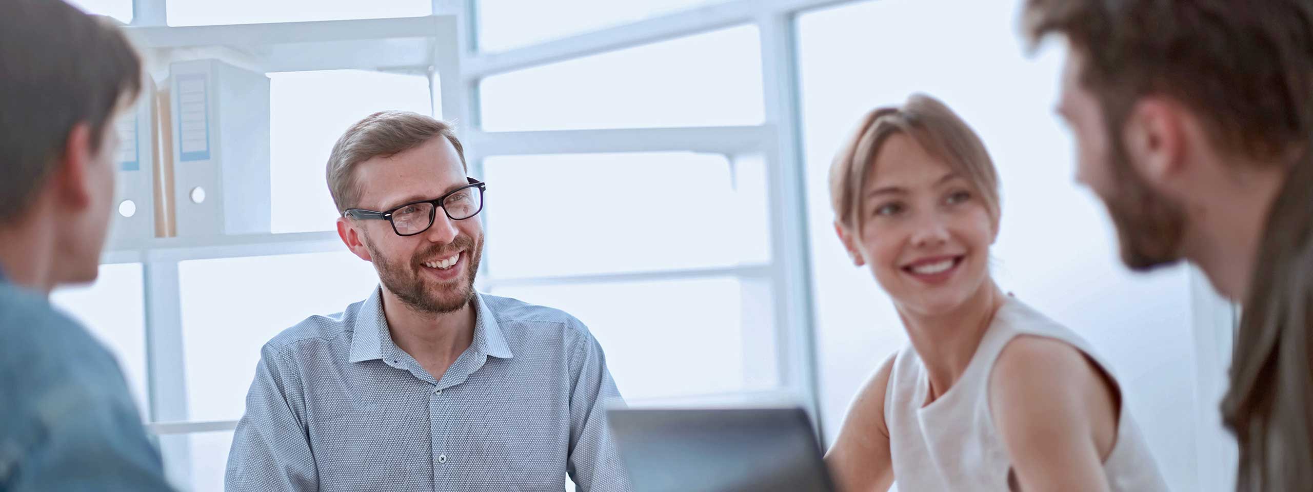 Vier Personen in einem Besprechungsraum diskutieren. Helle Umgebung mit Regalen und Dokumenten sichtbar.