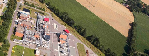 Eine Luftaufnahme einer Industrieanlage umgeben von Feldern und Bäumen. Rote Dächer und zahlreiche Rohre sind sichtbar.