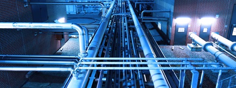 View of pipes in an industrial setting, illuminated by blue lights. A brick wall is visible in the background.
