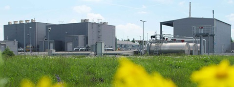 Industrielle Gebäude mit silberner Fassade und Rohren, vor einem grünen Feld mit Wildblumen unter einem blauen Himmel.