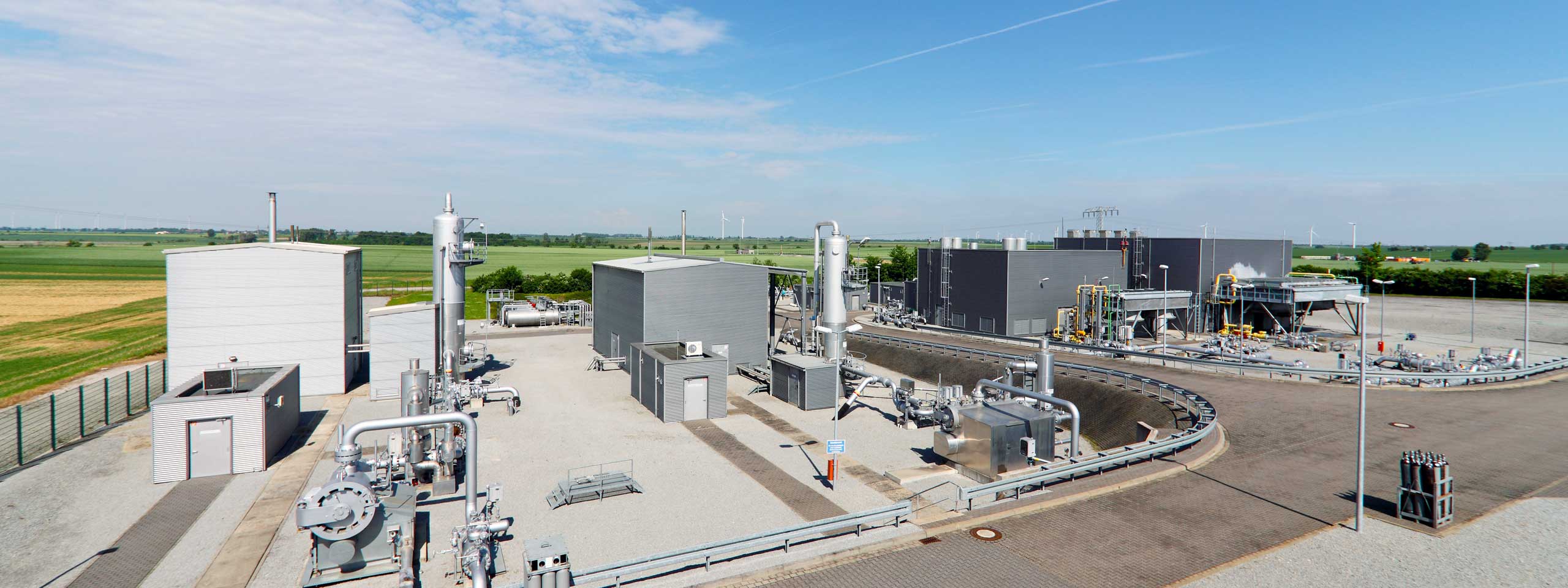 Industriegelände mit mehreren Gebäuden und Rohren. Im Hintergrund erstrecken sich grüne Felder und Windkraftanlagen.