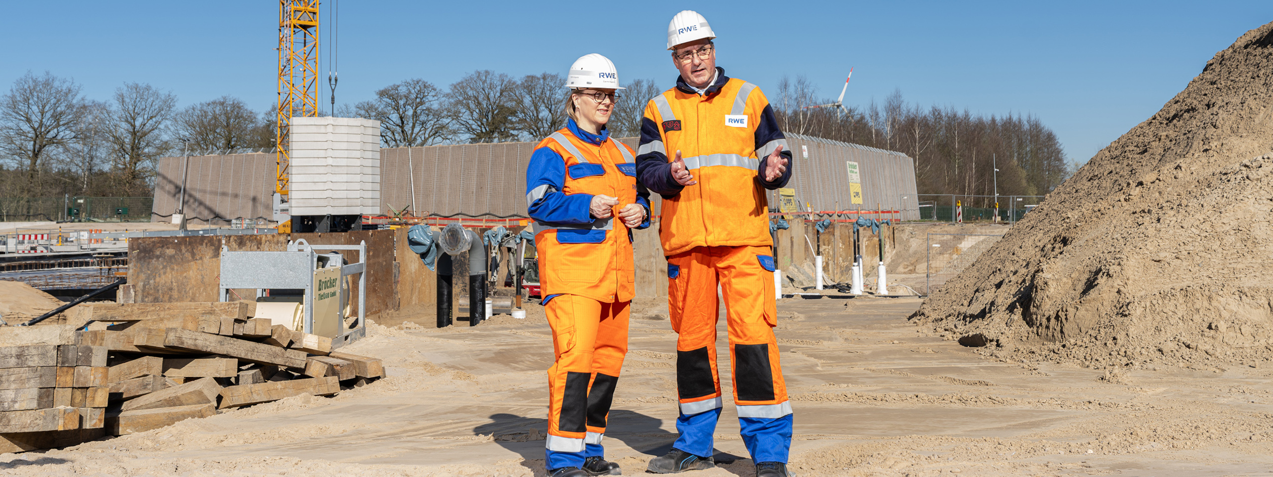Our management in orange and blue workwear stands on a construction site with equipment and machinery in the background.