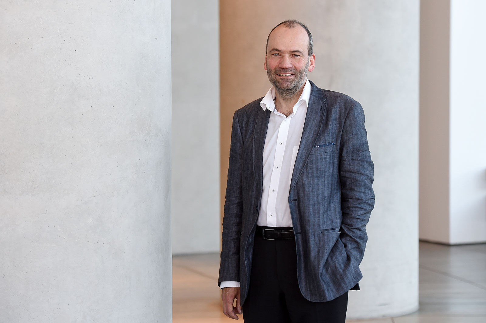 A person stands in front of a modern concrete wall, wearing a blazer and a white shirt. The background is minimalist.