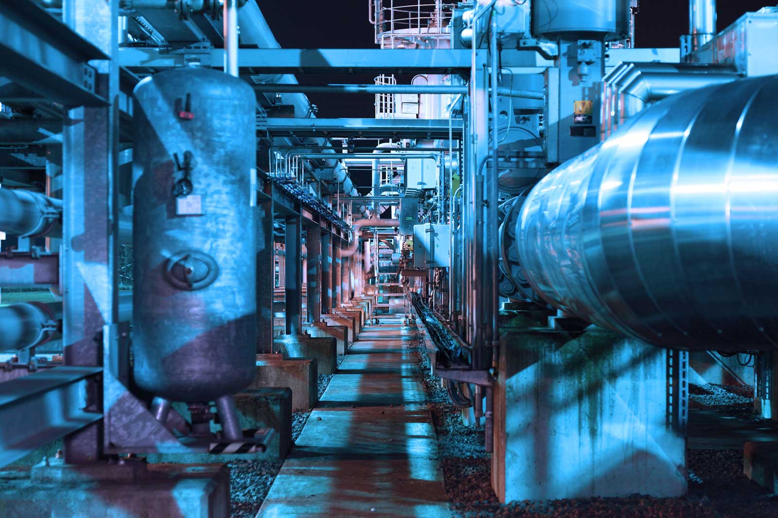 An industrial facility with pipes and tanks, illuminated by blue lighting at night.