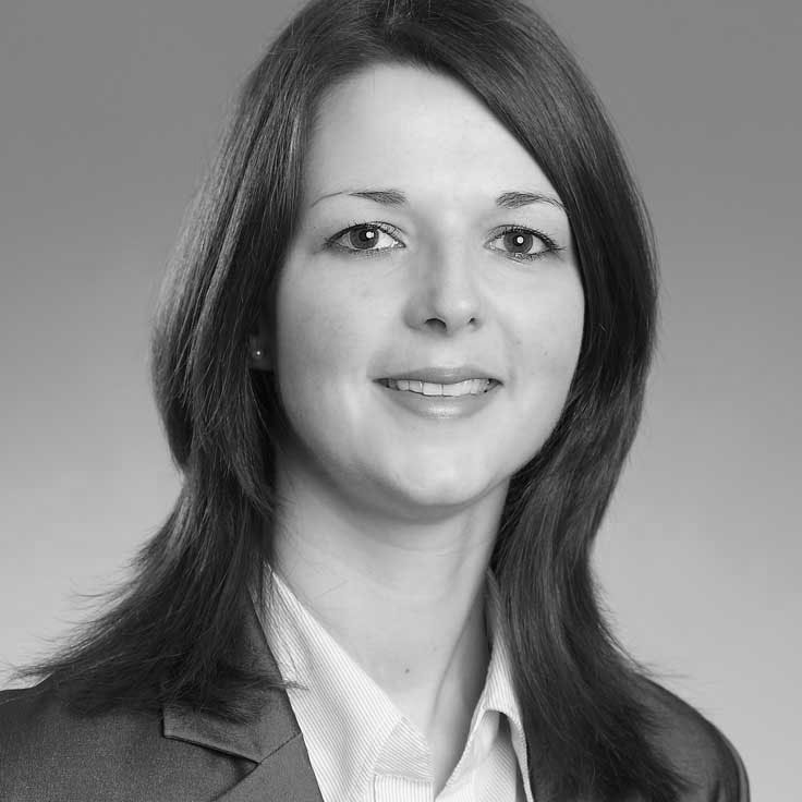 A person with long, straight hair wearing a grey blazer and a light shirt. The background is plain grey.