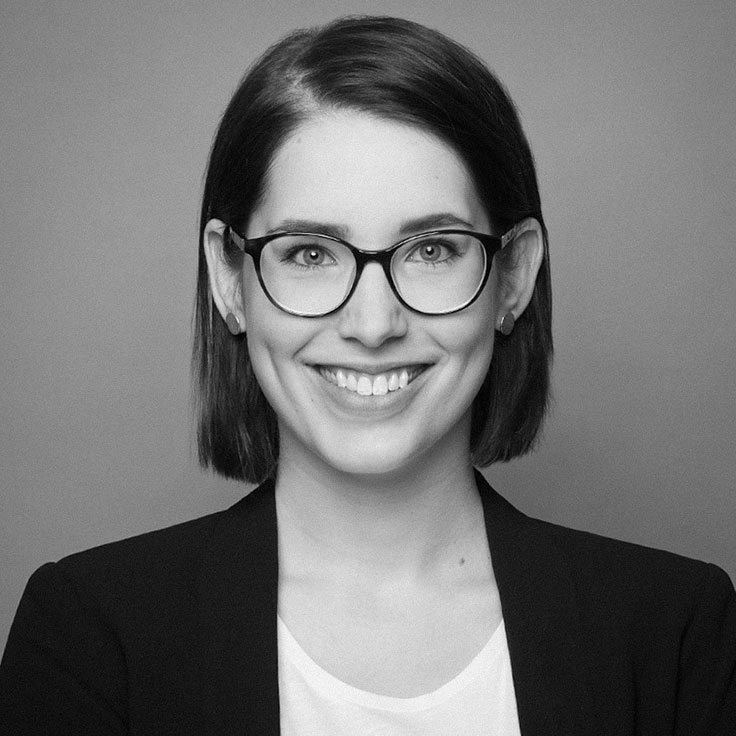 A person with short hair wears a black blazer and a white top. The background is grey.