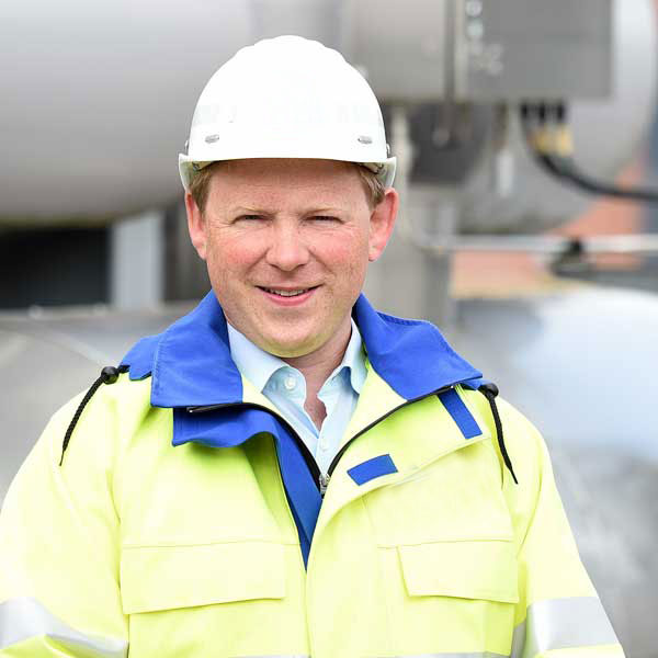 A worker in a yellow safety jacket with blue accents and a white helmet stands against an industrial backdrop.