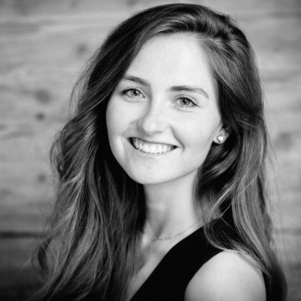 A person with long, curly hair wears a black top, with a wooden background.