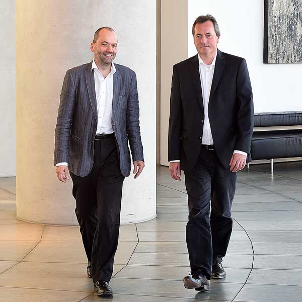 Two men in formal attire walk in a modern interior beside a column. The floor is bright and smooth.