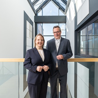 Two professionals in suits stand on a balcony with a modern architectural backdrop and large windows.